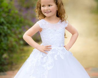 May include: A young girl in a white lace dress with a white headband. She is standing on a path with green foliage in the background.