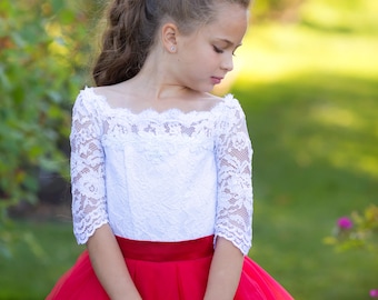 May include: A young girl wearing a white lace top with a red satin sash and a red tulle skirt. She is wearing a red flower in her hair and has a serious expression on her face.