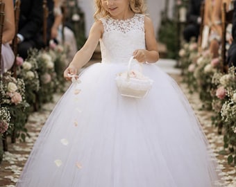 May include: A white floor-length dress with a sleeveless lace bodice and a full tulle skirt. The dress features a high neckline with floral lace detailing. A small white basket is held, and a floral crown is worn on the head with loose hair.