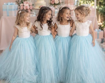 May include: Four children wearing matching floor-length dresses with white short-sleeved bodices and voluminous light blue tulle skirts. Each dress features a simple round neckline and a layered, flowing skirt design that reaches the floor.