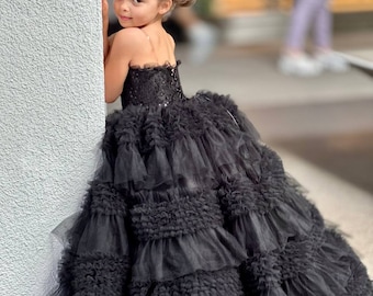 May include: A young girl wearing a black tulle dress with multiple layers of ruffles. The dress has a strapless bodice with a black lace overlay. The girl is standing next to a white wall and looking over her shoulder.