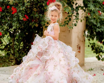 May include: A young girl wearing a pink and white floral gown with layered ruffles and a matching headband. The dress is adorned with delicate pink flowers and green leaves. The background features a lush green bush with red flowers.