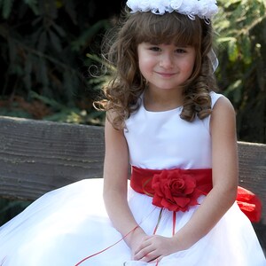 May include: A young girl wearing a white dress with a red sash and a flower crown. The dress has a red rose on the sash and is decorated with red rose petals.