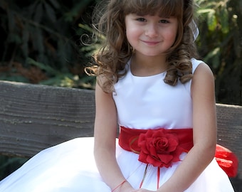 May include: A young girl wearing a white dress with a red sash and a flower crown. The dress has a red rose on the sash and is decorated with red rose petals.