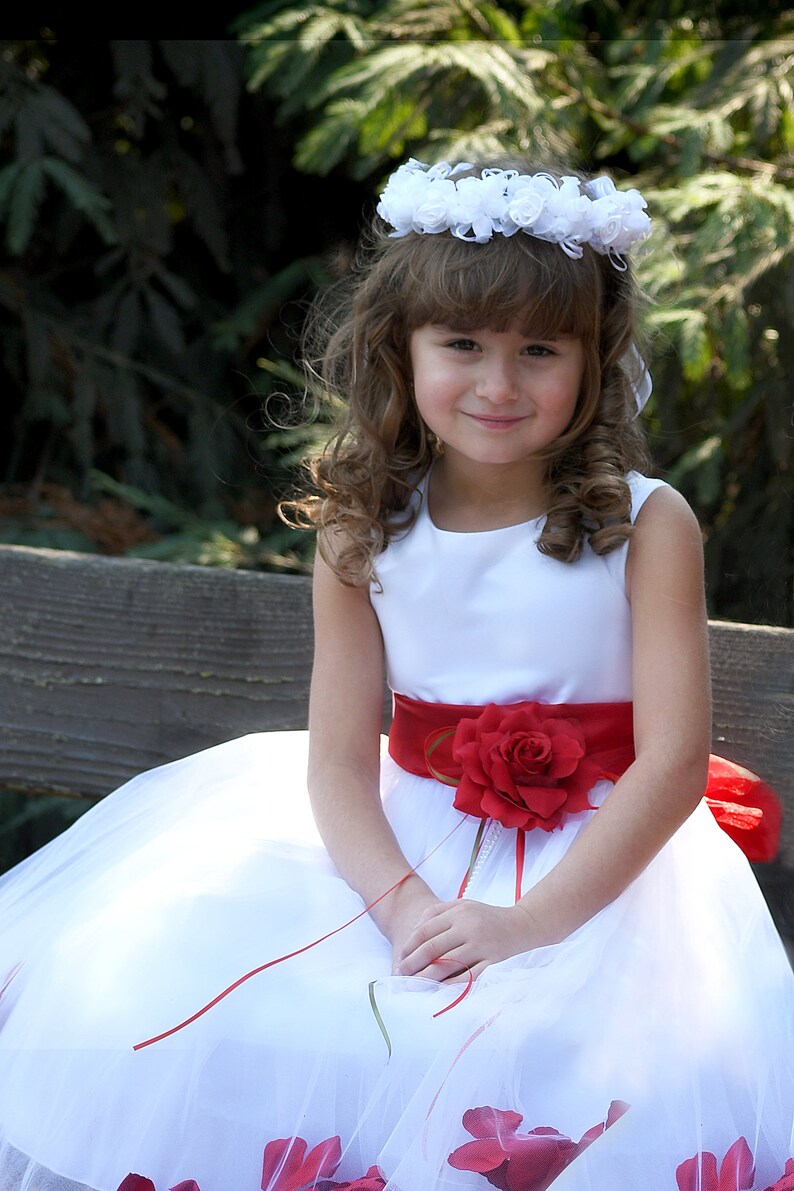 May include: A young girl wearing a white dress with a red sash and a flower crown. The dress has a red rose on the sash and is decorated with red rose petals.