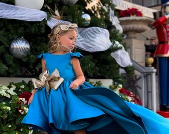 May include: A young girl wearing a teal blue high-low dress with a gold bow. The dress has a short front and a long train in the back. She is standing in front of a Christmas tree with ornaments and lights.