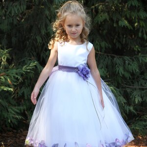 May include: A young girl wearing a white tulle dress with a purple sash and flower accents. The dress has a layer of purple flower petals around the hem.