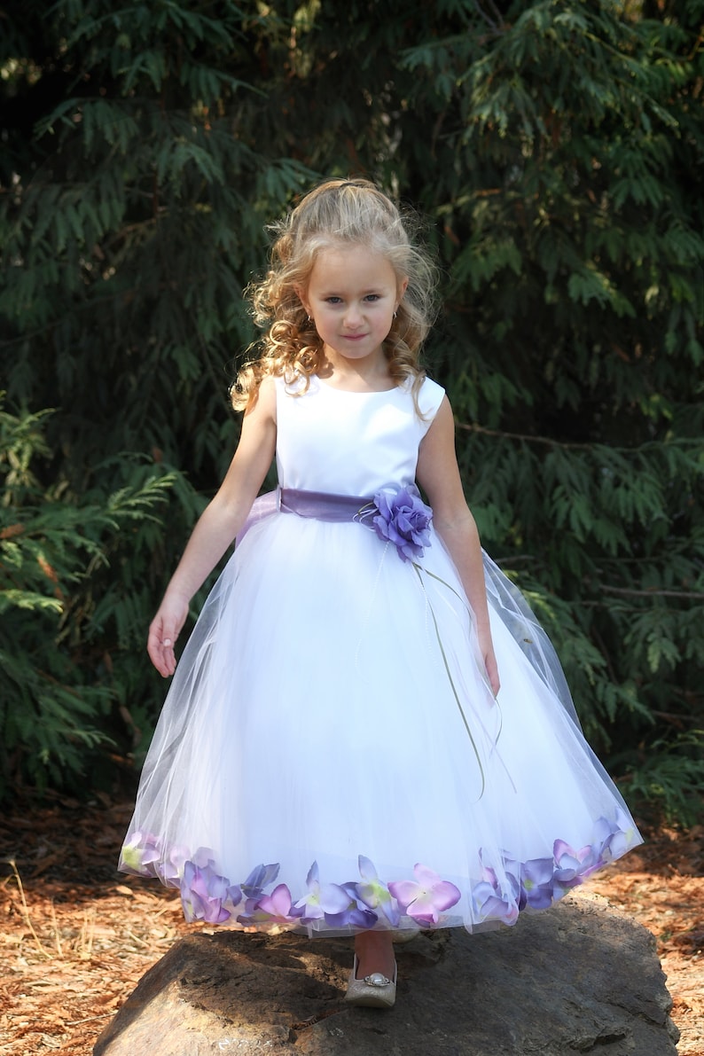 May include: A young girl wearing a white tulle dress with a purple sash and flower accents. The dress has a layer of purple flower petals around the hem.