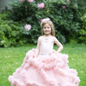 Puede incluir: Una ni&ntilde;a peque&ntilde;a con un vestido de tul rosa con m&uacute;ltiples capas de volantes. Est&aacute; de pie en un &aacute;rea cubierta de hierba con vegetaci&oacute;n verde en el fondo.