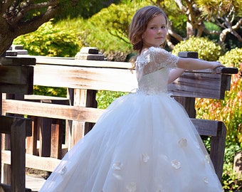 May include: A young girl in a white tulle dress with lace sleeves and flower embellishments poses on a wooden bridge in a garden setting.