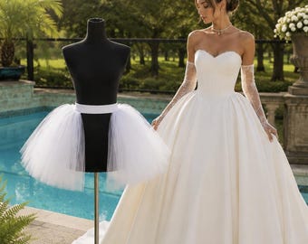 May include: An open front bridal petticoat with a white waistband and layered tulle, displayed on a black mannequin. The text reads: OPEN FRONT BRIDAL PETTICOAT, VOLUME AT SIDES & BACK – SMOOTH IN FRONT. A white gown is shown for comparison.