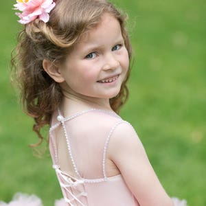 Puede incluir: Una ni&ntilde;a peque&ntilde;a con un vestido de tul rosa con una espalda de encaje blanco. Lleva un accesorio para el pelo con una flor rosa y una pluma. Est&aacute; sonriendo y mirando por encima del hombro.