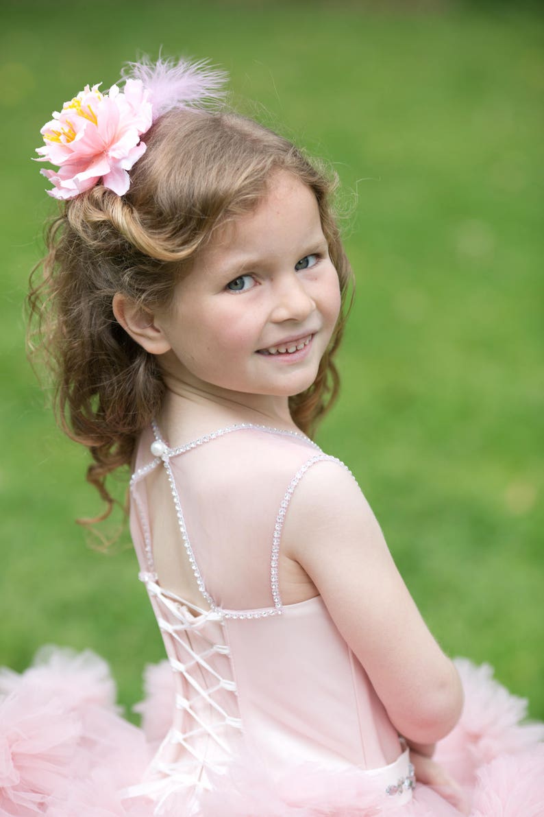 Puede incluir: Una ni&ntilde;a peque&ntilde;a con un vestido de tul rosa con una espalda de encaje blanco. Lleva un accesorio para el pelo con una flor rosa y una pluma. Est&aacute; sonriendo y mirando por encima del hombro.