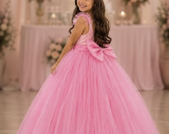 May include: A floor-length pink dress featuring a sleeveless bodice with ruffled shoulder straps, a full tulle skirt, and a large satin bow at the waist. The dress is paired with a silver-toned tiara worn on long, dark, wavy hair.