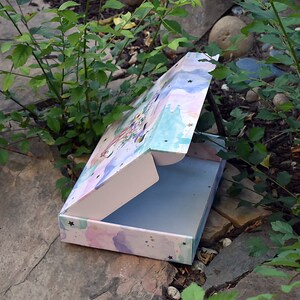 Pu&ograve; includere: Una scatola aperta con un design ad acquerello rosa, blu e verde. La scatola ha stelle dorate ed &egrave; decorata con un motivo floreale fantasioso. La scatola &egrave; aperta e l'interno &egrave; vuoto.