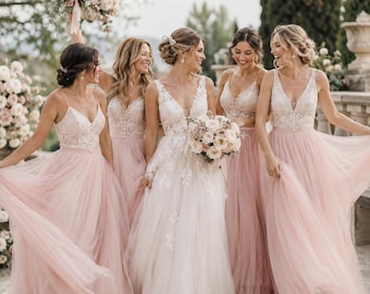 May include: A group of five dresses featuring lace bodices with V-necklines and flowing tulle skirts. Four dresses are in a soft blush pink, while the central dress is white with floral lace appliqués. Each dress has thin straps.