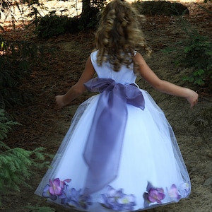 May include: A young girl wearing a white dress with a purple sash and floral accents. She is wearing sparkly silver shoes and has her hair pulled back.