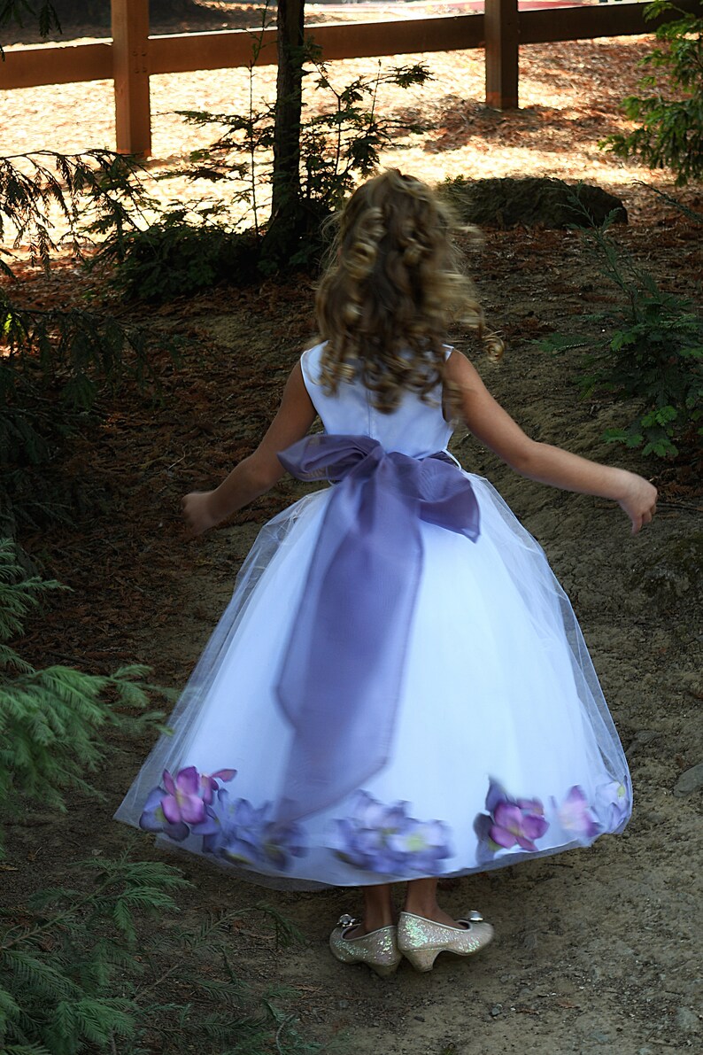 May include: A young girl wearing a white dress with a purple sash and floral accents. She is wearing sparkly silver shoes and has her hair pulled back.