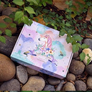 Pu&ograve; includere: Una scatola regalo rosa e viola con un design ad acquerello che presenta un unicorno e fiori. La scatola ha una scritta dorata che recita "Fairy Unicorn" e "Noble Qualities, Kind and Pure".
