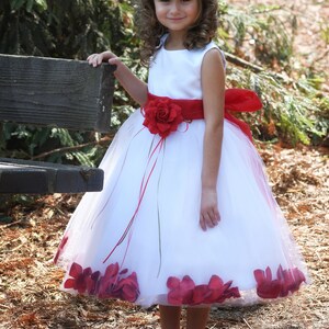 May include: A young girl wearing a white dress with a red sash and a flower accent. The dress has a tulle skirt with red flower petals around the hem. She is wearing a white flower crown and white shoes.