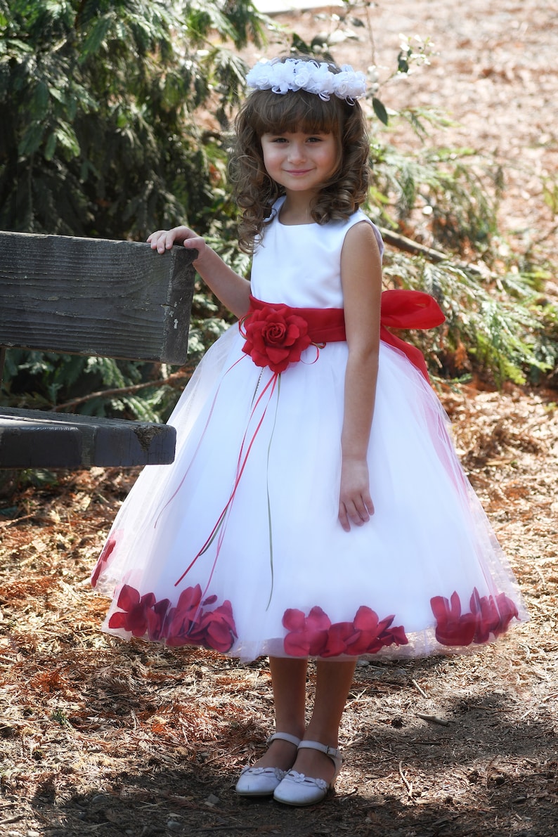 May include: A young girl wearing a white dress with a red sash and a flower accent. The dress has a tulle skirt with red flower petals around the hem. She is wearing a white flower crown and white shoes.