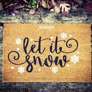 Puede incluir: Una alfombra de coco marr&oacute;n con el texto "Let it snow" en letras cursivas negras y detalles de copos de nieve blancos. El texto "KK Designs" est&aacute; impreso en la esquina superior derecha.