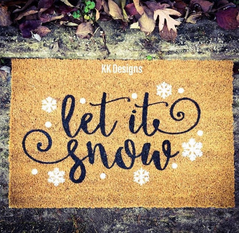 Puede incluir: Una alfombra de coco marr&oacute;n con el texto "Let it snow" en letras cursivas negras y detalles de copos de nieve blancos. El texto "KK Designs" est&aacute; impreso en la esquina superior derecha.