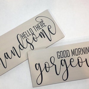 May include: Two rectangular wooden signs with light beige backgrounds. One sign reads "HELLO THERE handsome" in black cursive and block letters. The other sign says "GOOD MORNIN' gorgeous" in black cursive and block letters.