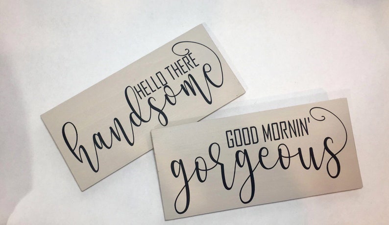 May include: Two rectangular wooden signs with light beige backgrounds. One sign reads "HELLO THERE handsome" in black cursive and block letters. The other sign says "GOOD MORNIN' gorgeous" in black cursive and block letters.