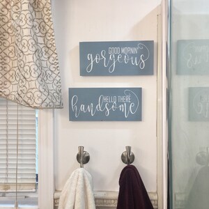 May include: Two rectangular, light blue wooden signs with white cursive text that reads "Good Mornin' Gorgeous" and "Hello There Handsome" are mounted on a white wall. Below, two metal hooks hold a white and a burgundy towel. A window with blinds is on the left.