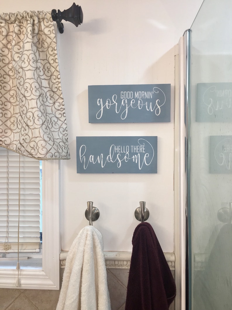 May include: Two rectangular, light blue wooden signs with white cursive text that reads "Good Mornin' Gorgeous" and "Hello There Handsome" are mounted on a white wall. Below, two metal hooks hold a white and a burgundy towel. A window with blinds is on the left.