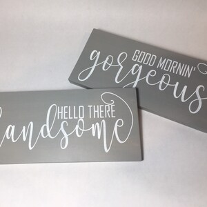 May include: Two rectangular gray wooden signs with white cursive text. One sign reads "Hello There Handsome" and the other reads "Good Mornin' Gorgeous". The signs are likely for home decor.