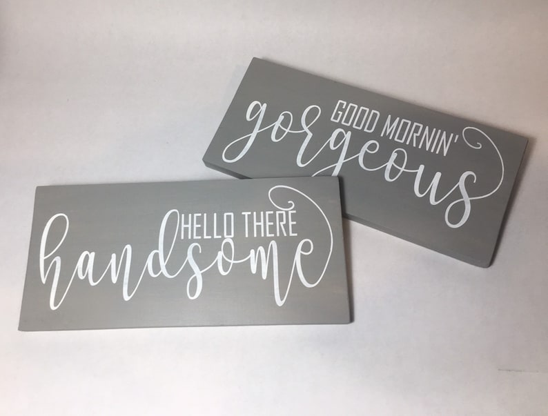 May include: Two rectangular gray wooden signs with white cursive text. One sign reads "Hello There Handsome" and the other reads "Good Mornin' Gorgeous". The signs are likely for home decor.