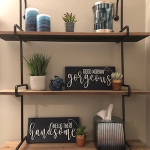 May include: A decorative wall shelf with two wooden shelves and black metal supports. The shelves hold various decorative items, including candles, potted plants, and signs with the phrases "Good Mornin' Gorgeous" and "Hello There Handsome."