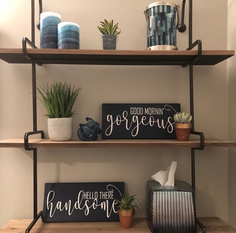 May include: A decorative wall shelf with two wooden shelves and black metal supports. The shelves hold various decorative items, including candles, potted plants, and signs with the phrases "Good Mornin' Gorgeous" and "Hello There Handsome."