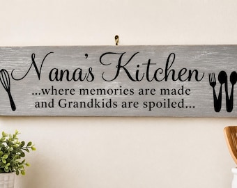 May include: A rectangular gray wooden sign with a distressed finish featuring the black text Nana's Kitchen ...where memories are made and Grandkids are spoiled... flanked by a black whisk icon on the left and a fork and spoon icon on the right.