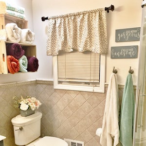 May include: A bathroom scene featuring a white toilet, a window with a patterned valance, and two wooden crates holding rolled towels. Two signs read "Good Morning Gorgeous" and "Hello There Handsome". Two towels hang from hooks.