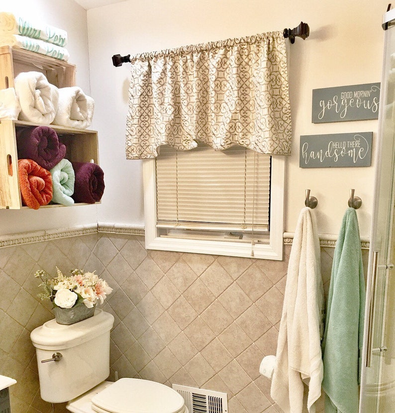 May include: A bathroom scene featuring a white toilet, a window with a patterned valance, and two wooden crates holding rolled towels. Two signs read "Good Morning Gorgeous" and "Hello There Handsome". Two towels hang from hooks.