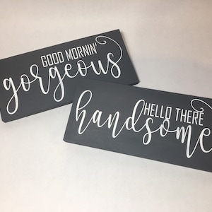 May include: Two rectangular, dark gray wooden signs with white cursive text. One sign reads "GOOD MORNING gorgeous" and the other says "HELLO THERE handsome". The signs are leaning against each other.