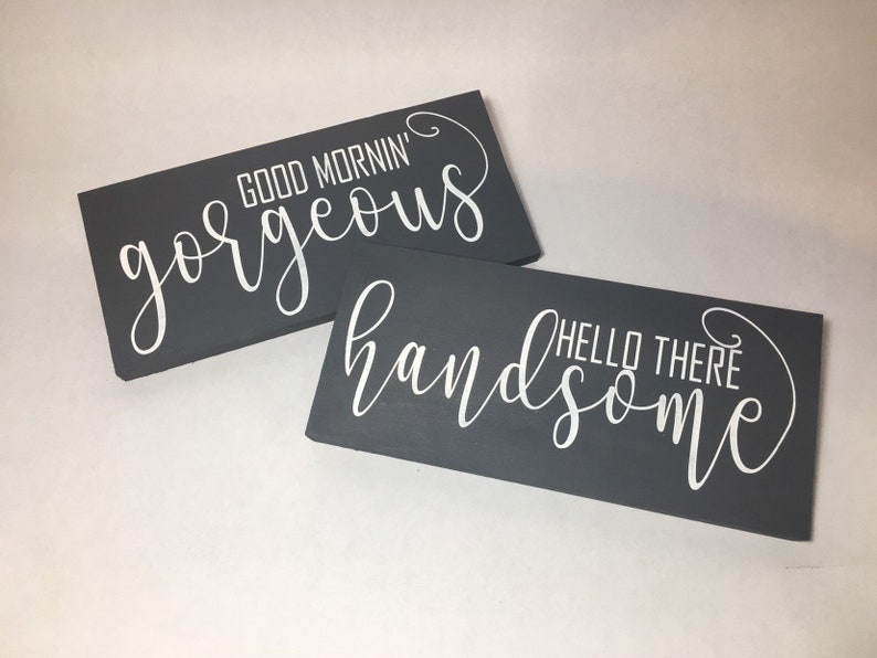 May include: Two rectangular, dark gray wooden signs with white cursive text. One sign reads "GOOD MORNING gorgeous" and the other says "HELLO THERE handsome". The signs are leaning against each other.