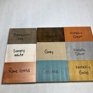May include: Nine wooden color swatches labeled with paint names like "Barnwood Gray," "Walnut," and "Metallic Copper." Other colors include "Simply White," "Grey," "Rose Gold," "Cascade," "Metallic Silver," and "Metallic Gold." The colors are painted on small wooden boards.