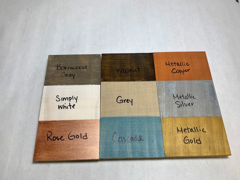 May include: Nine wooden color swatches labeled with paint names like "Barnwood Gray," "Walnut," and "Metallic Copper." Other colors include "Simply White," "Grey," "Rose Gold," "Cascade," "Metallic Silver," and "Metallic Gold." The colors are painted on small wooden boards.