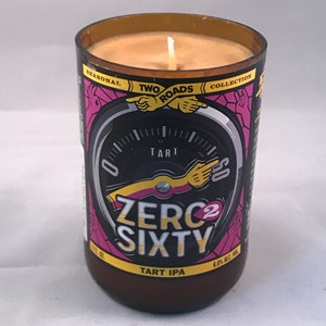 May include: A brown glass candle jar with a yellow and black label. The label features a speedometer with the text "Two Roads Collection" and "Zero2Sixty Tart IPA".