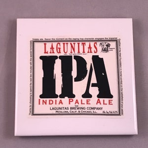 May include: A white ceramic coaster with a red and black illustration of a beer label. The label reads "Lagunitas IPA India Pale Ale" and features a cartoon dog with the text "Doggone Good".
