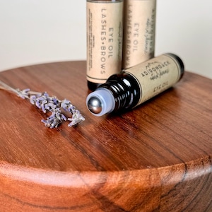 May include: Three brown glass roller bottles with labels that read "Eye Oil Lashes & Brows" and "Adirondack Wild Ferns Eye Oil". The bottles are on a wooden surface with a sprig of lavender.