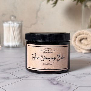 May include: A jar of Tallow Cleansing Balm with a black lid and a label that reads "Adirondack natural skincare". The label also says "Charcoal" and "6 oz / 171 gm". The jar is on a white and gray tiled surface.