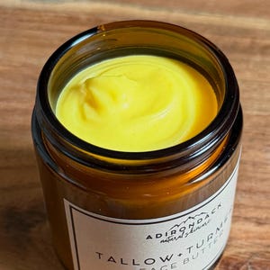 May include: An amber glass jar filled with yellow face butter. The jar has a black lid and a white label with black text. The label reads "ADIRONDACK natural skincare" and "TALLOW + TURMERIC FACE BUTTER 100 g". The butter has a creamy, smooth texture.