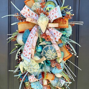 May include: A decorative wreath with a coastal theme, featuring a large bow with seashell patterned ribbon, burlap accents, and various blue and teal ornaments. The wreath is adorned with faux greenery and white accents, creating a textured and layered design.