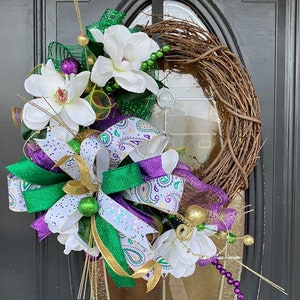 May include: A green, purple, and gold wreath with white magnolia flowers, a bow, and a paisley pattern. The wreath is made of twigs and is decorated with beads and other embellishments.