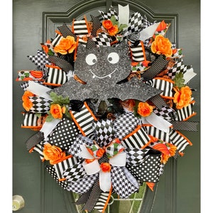 May include: A black and white Halloween wreath with a black bat in the center. The wreath is decorated with orange and white ribbon, black and white polka dot ribbon, and black and white checkered ribbon. There are also orange flowers around the wreath.